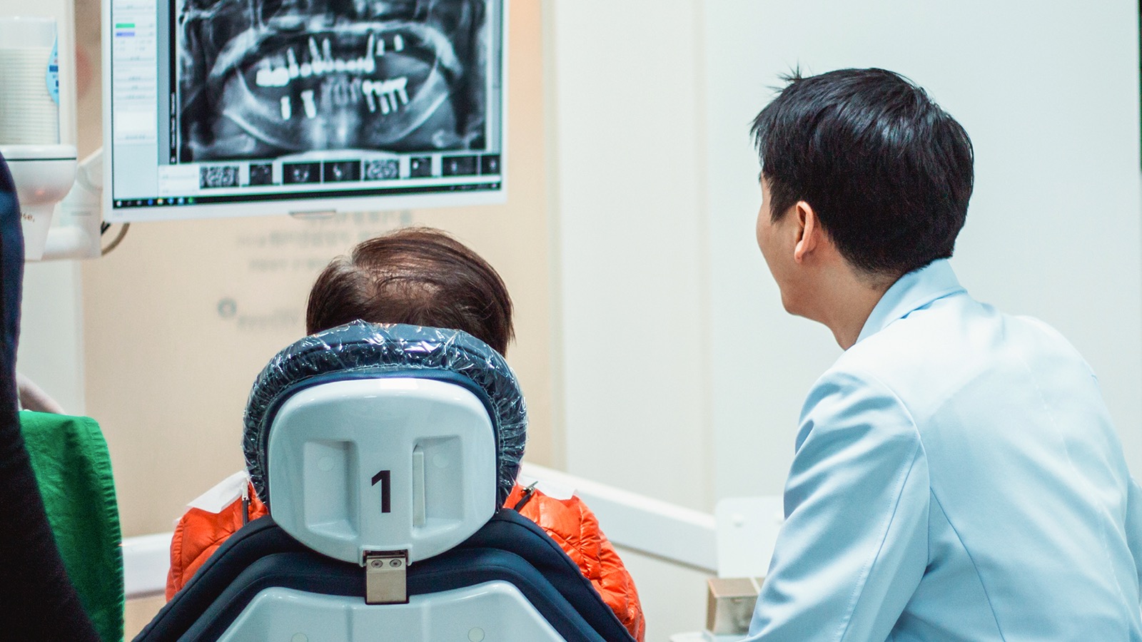 치과의사와 진료 의자에 앉은 환자가 임플란트가 여러 개 보이는 파노라마 엑스레이 화면을 함께 보는 모습